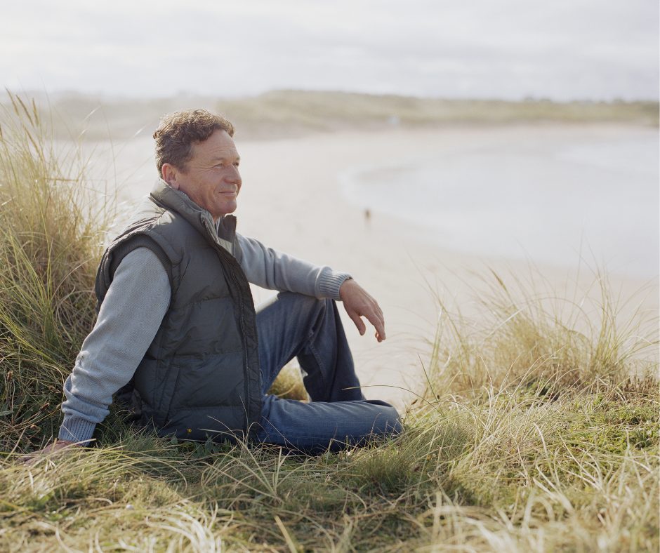 man in his fifties sitting in coastal dune grass looking out over the beach reflecting on direction in the second half of life