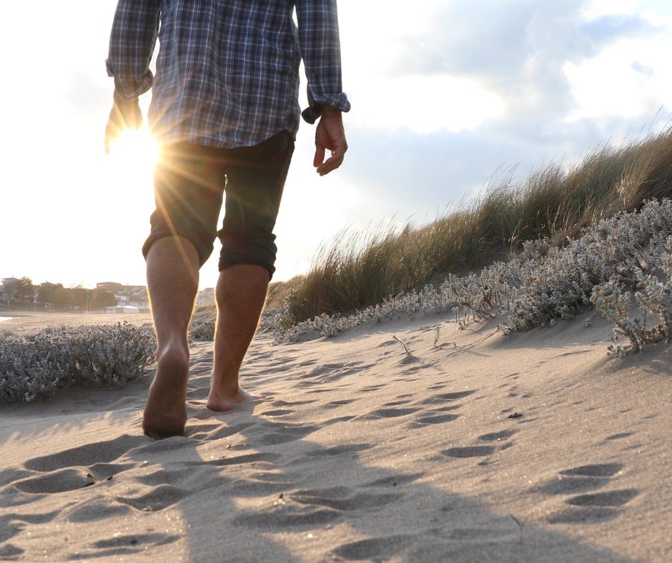 Person walking barefoot on a sandy beach at sunset, leaving footprints with dune grass on the right.